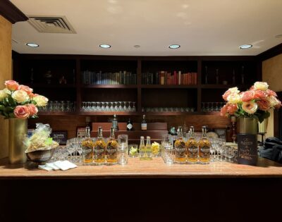 image of a bar setup with pink and white roses in a vases on either side, bottles of liquor and glasses on counter, with a bowl of wrapped treats on the left side. Back bar is dark wood with glasses in shelves
