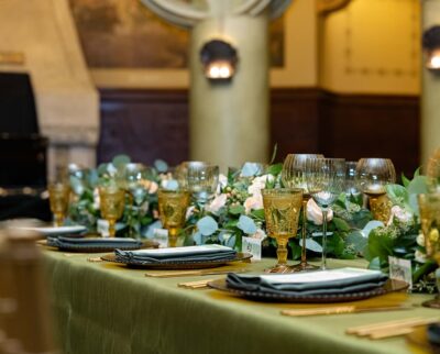 Image of a table scape at a holiday party, with green and gold glassware, gold flatware, and dark peacock green napkins. small menus are laid on top of the folded linens.